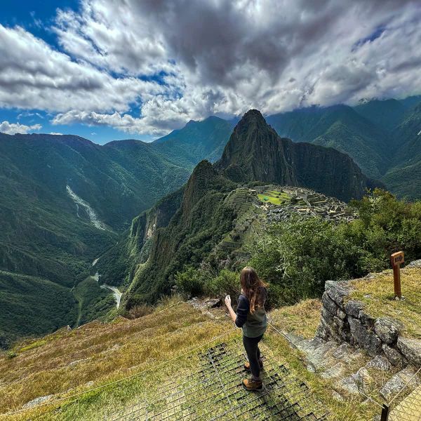 Person standing on a grassy terrace overlooking Machu Picchu, surrounded by mountains under a cloudy sky.