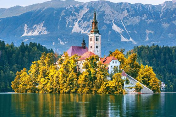Scenic view of an island with a church and autumn trees on a tranquil lake, surrounded by mountains under a clear sky.