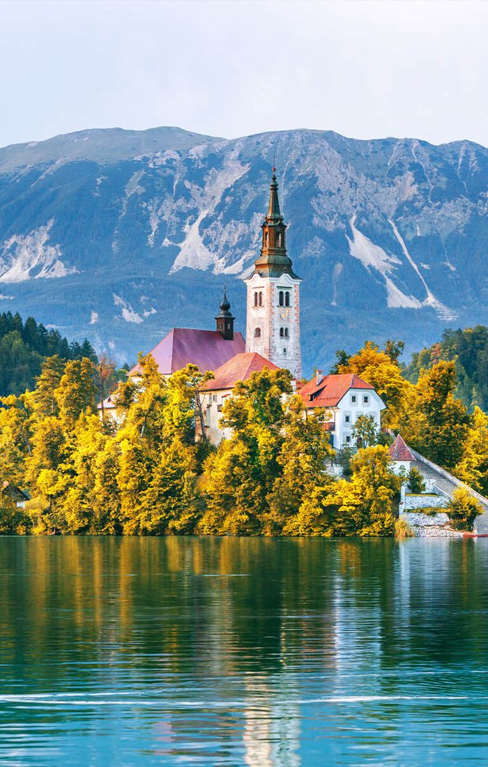 Scenic view of an island with a church and autumn trees on a tranquil lake, surrounded by mountains under a clear sky.