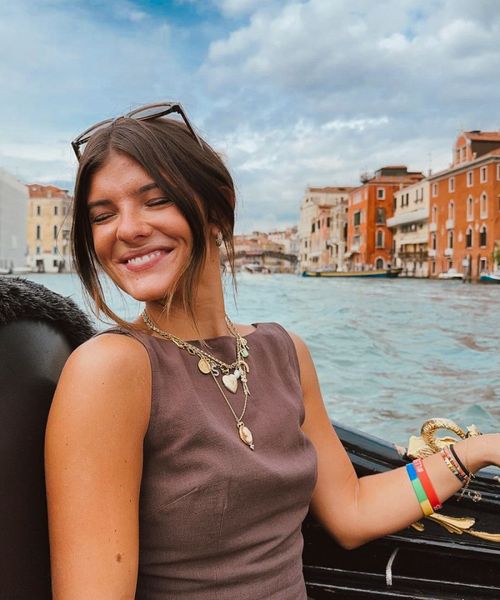 Smiling woman with sunglasses on her head, seated on a gondola in Venice, with colorful buildings and a canal in the background.