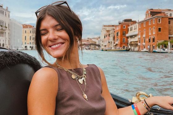 Smiling woman with sunglasses on her head, seated on a gondola in Venice, with colorful buildings and a canal in the background.