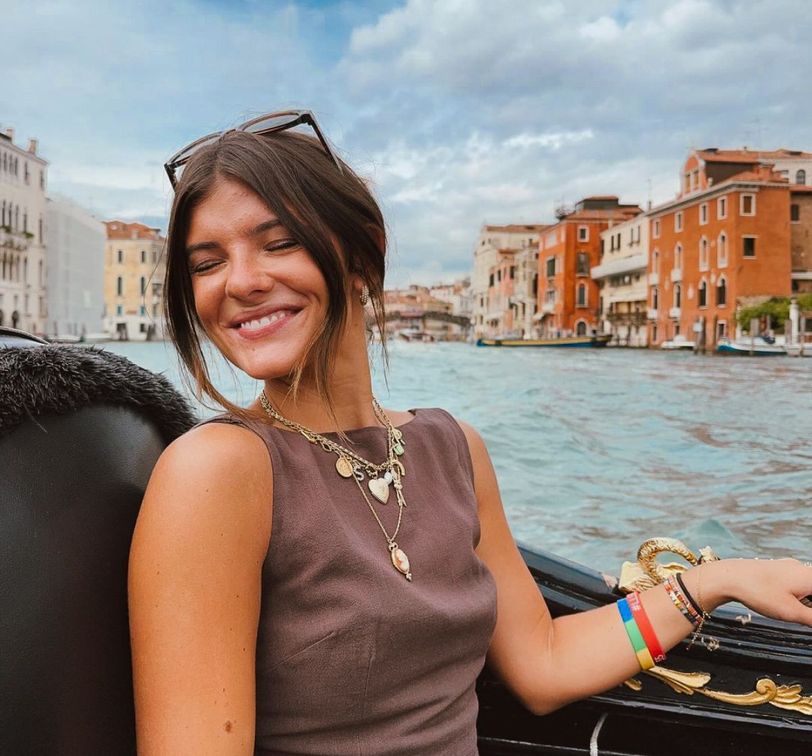 Smiling woman with sunglasses on her head, seated on a gondola in Venice, with colorful buildings and a canal in the background.