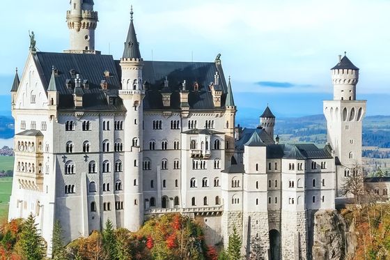 A majestic castle with numerous towers and spires stands atop a forested hill, under a clear blue sky.