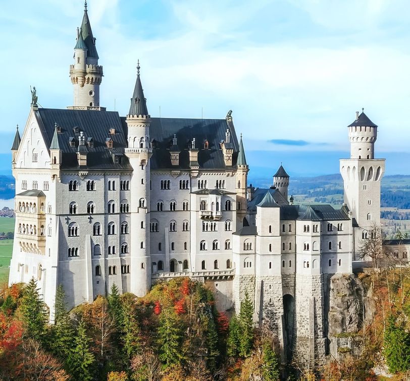 A majestic castle with numerous towers and spires stands atop a forested hill, under a clear blue sky.
