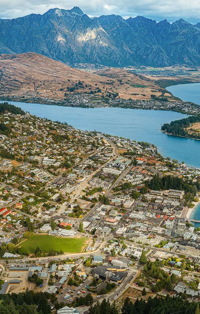 Aerial view of a town nestled between a lake and mountains, with scattered buildings, roads, and lush greenery under a partly cloudy sky.