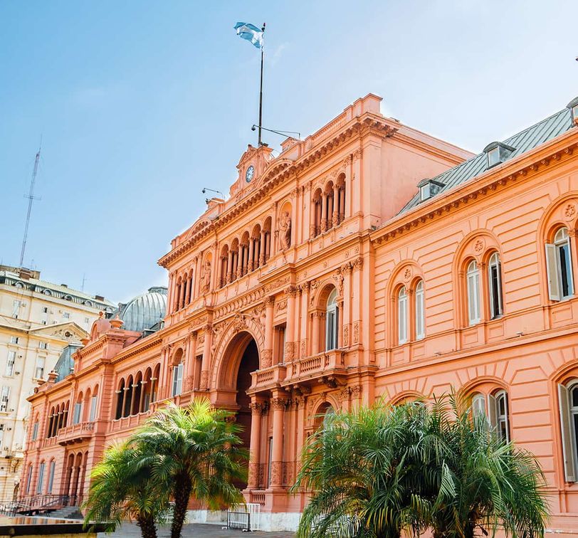 Historic pink building with ornate architecture, palm trees in front, and an Argentine flag on top, under a clear blue sky.