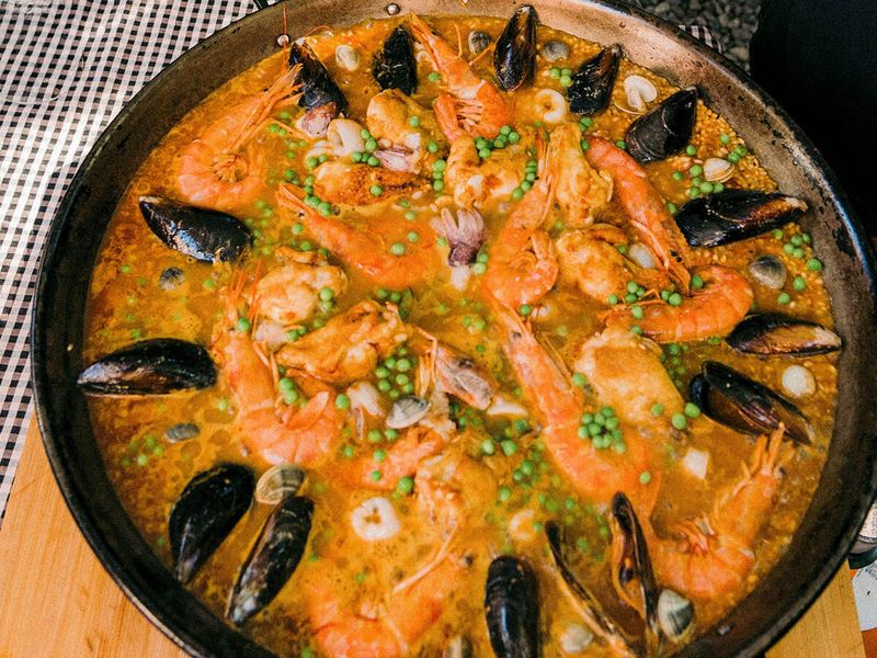 A large pan of seafood paella with shrimp, mussels, peas, and rice on a wooden board, garnished and ready to serve.