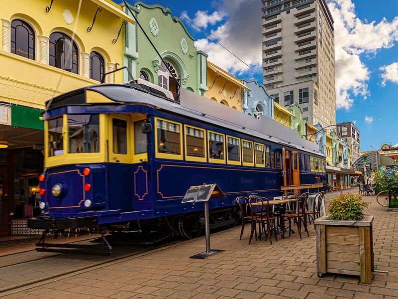A vintage blue and yellow tram travels down a pedestrian street with colorful buildings and outdoor seating, under a partly cloudy sky.