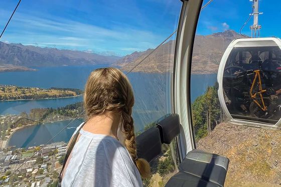 A person with braided hair sits inside a gondola, overlooking a scenic view of a lake, mountains, and a town under a clear blue sky.