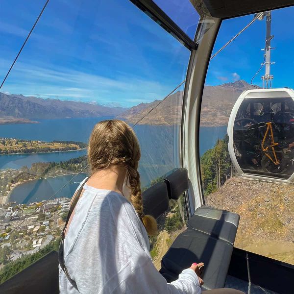 A person with braided hair sits inside a gondola, overlooking a scenic view of a lake, mountains, and a town under a clear blue sky.