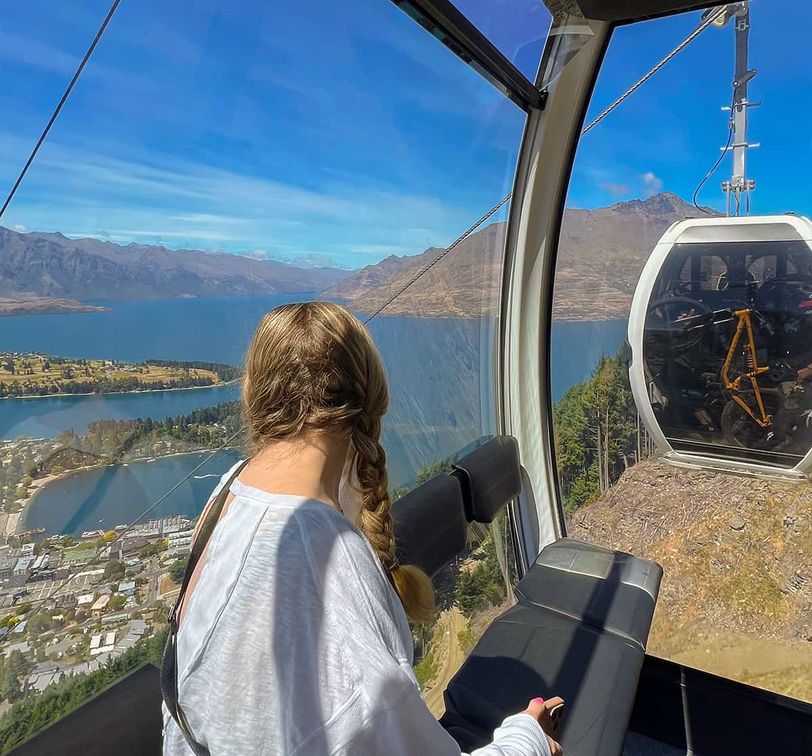 A person with braided hair sits inside a gondola, overlooking a scenic view of a lake, mountains, and a town under a clear blue sky.