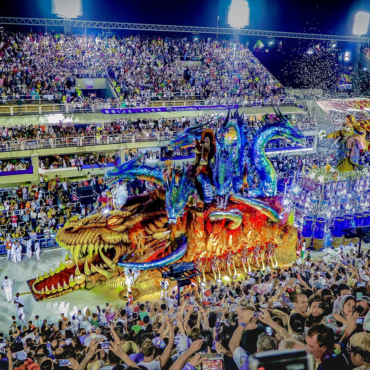 Colorful parade float with dragon design at a crowded carnival, surrounded by cheering spectators in a brightly lit stadium.