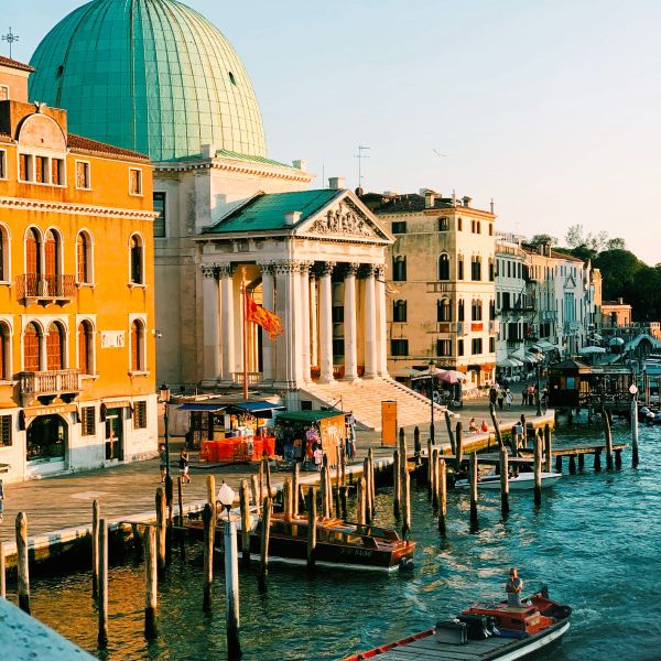 View of Venice canal with moored boats, historic buildings, and a domed church bathed in warm sunlight.