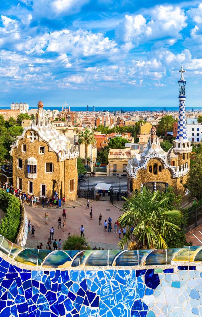 Colorful view of Parc Güell in Barcelona, featuring vibrant mosaic tiles and unique, whimsical buildings under a bright, cloudy sky.