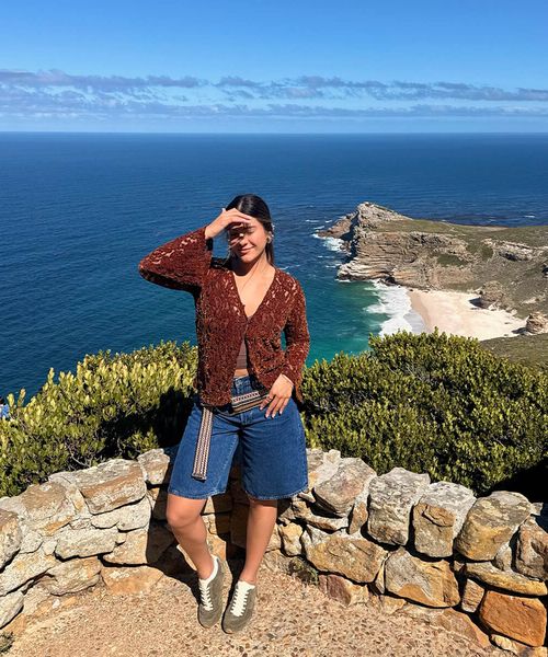 Woman posing on a stone wall with a stunning coastal view in the background; clear blue sky and ocean visible.