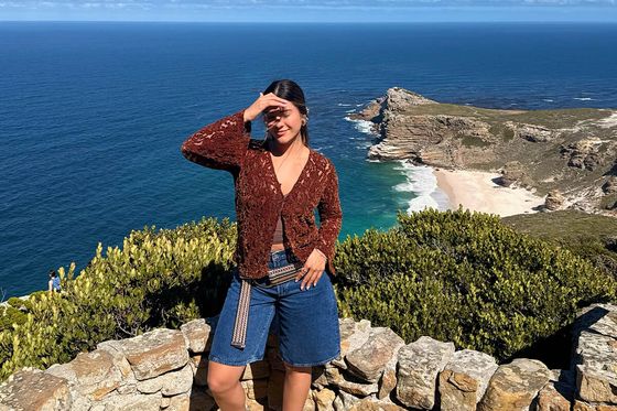 Woman posing on a stone wall with a stunning coastal view in the background; clear blue sky and ocean visible.