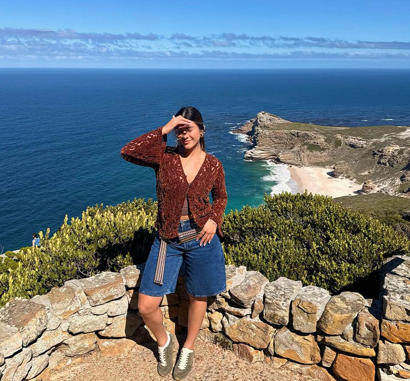 Woman posing on a stone wall with a stunning coastal view in the background; clear blue sky and ocean visible.