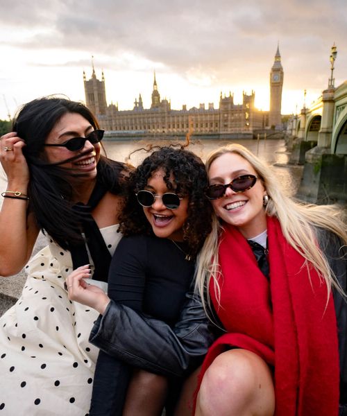 Three women in sunglasses smile and pose near the River Thames with the Houses of Parliament and Big Ben in the background at sunset.