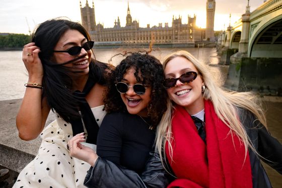 Three women in sunglasses smile and pose near the River Thames with the Houses of Parliament and Big Ben in the background at sunset.