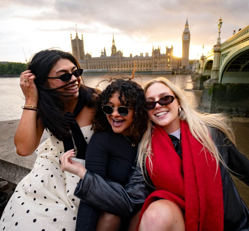 Three women in sunglasses smile and pose near the River Thames with the Houses of Parliament and Big Ben in the background at sunset.