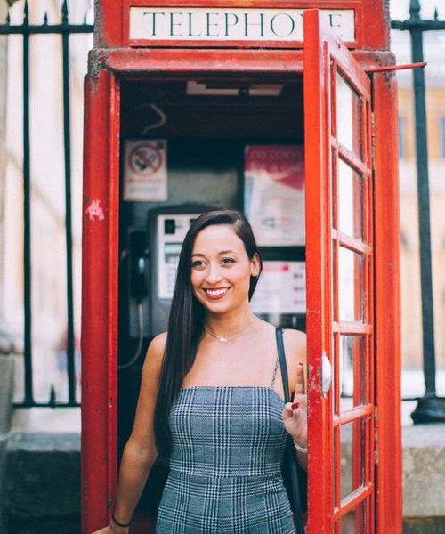 A woman smiling inside a traditional red British telephone booth, wearing a checkered dress, with part of her hand on the door.