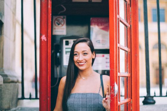 A woman smiling inside a traditional red British telephone booth, wearing a checkered dress, with part of her hand on the door.