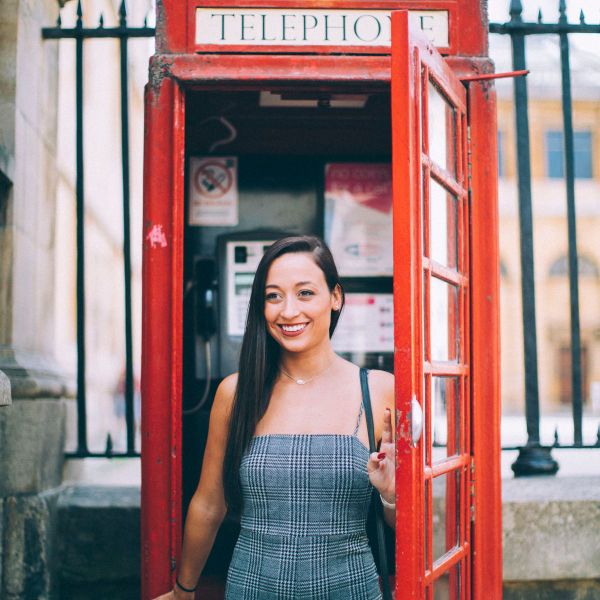 A woman smiling inside a traditional red British telephone booth, wearing a checkered dress, with part of her hand on the door.