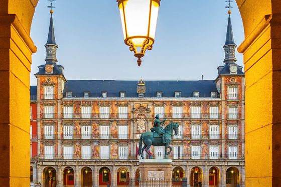 Arched view of Plaza Mayor, Madrid at dawn, showcasing a central equestrian statue and ornate building facade under a glowing lantern.