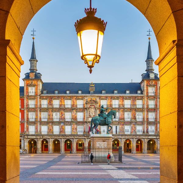 Arched view of Plaza Mayor, Madrid at dawn, showcasing a central equestrian statue and ornate building facade under a glowing lantern.