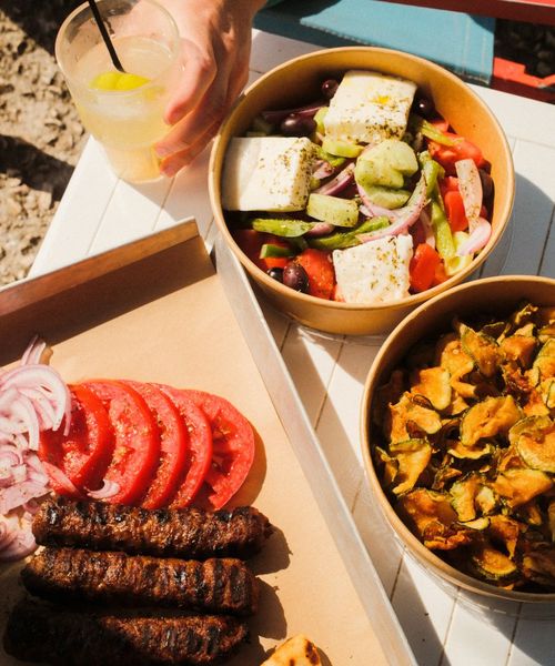 Grilled sausages, sliced tomatoes, and onions on a tray, with bowls of Greek salad and fried zucchini slices, next to a drink with a lime wedge.
