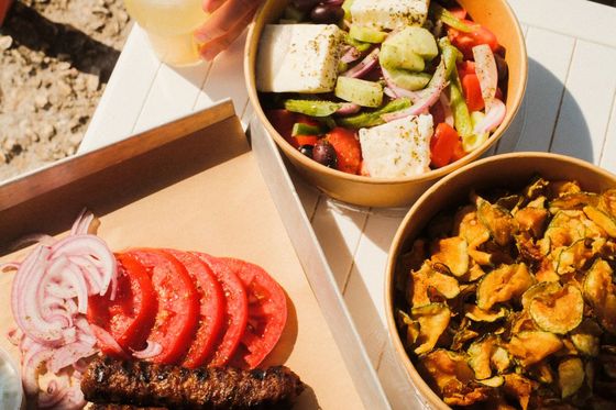 Grilled sausages, sliced tomatoes, and onions on a tray, with bowls of Greek salad and fried zucchini slices, next to a drink with a lime wedge.
