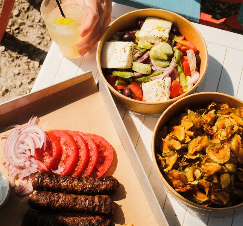 Grilled sausages, sliced tomatoes, and onions on a tray, with bowls of Greek salad and fried zucchini slices, next to a drink with a lime wedge.