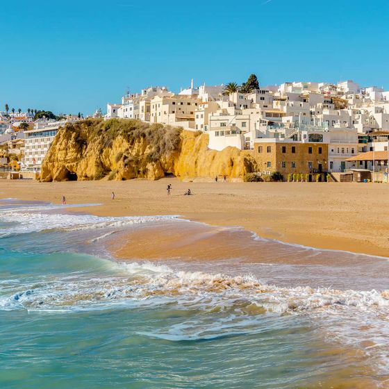 Golden sandy beach with gentle waves, coastal cliff topped with white buildings under a clear blue sky. A few people stroll along the shore.
