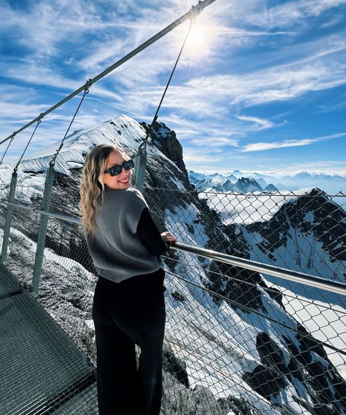 A woman in sunglasses smiles on a snowy mountain walkway, with a stunning view of sunlit peaks and a blue sky in the background.