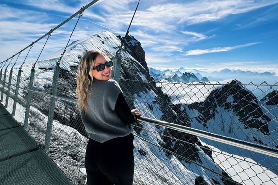 A woman in sunglasses smiles on a snowy mountain walkway, with a stunning view of sunlit peaks and a blue sky in the background.