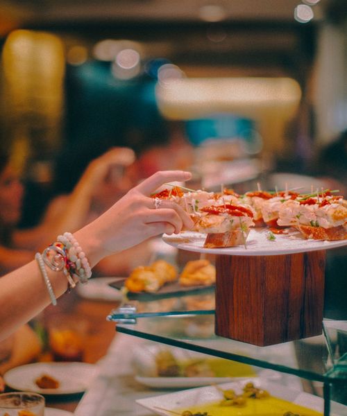 A person reaches for a piece of finger food on a decorative platter in a restaurant setting, with a blurred background of diners.