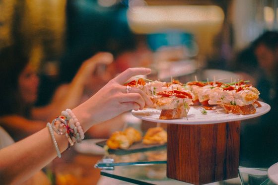 A person reaches for a piece of finger food on a decorative platter in a restaurant setting, with a blurred background of diners.