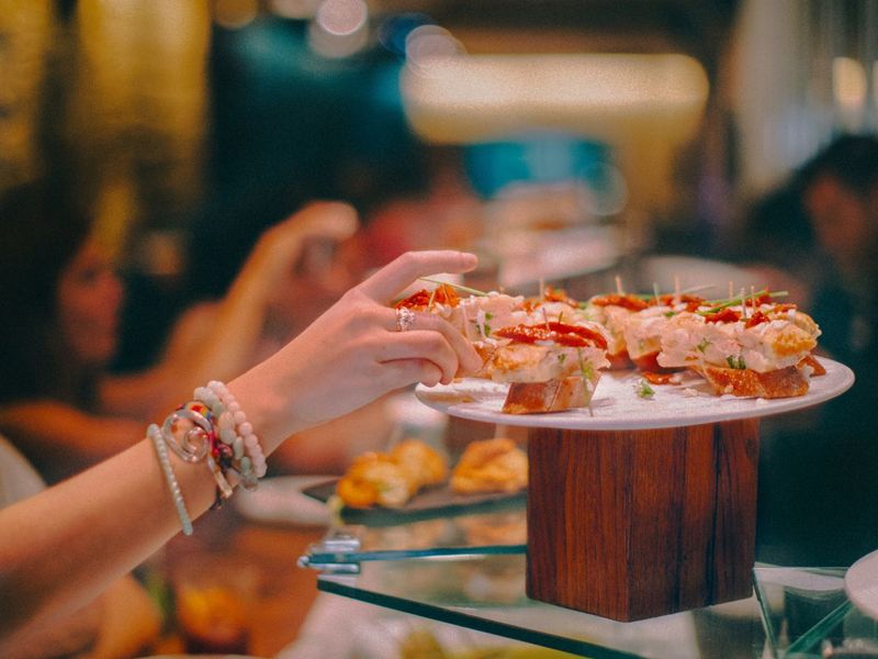 A person reaches for a piece of finger food on a decorative platter in a restaurant setting, with a blurred background of diners.