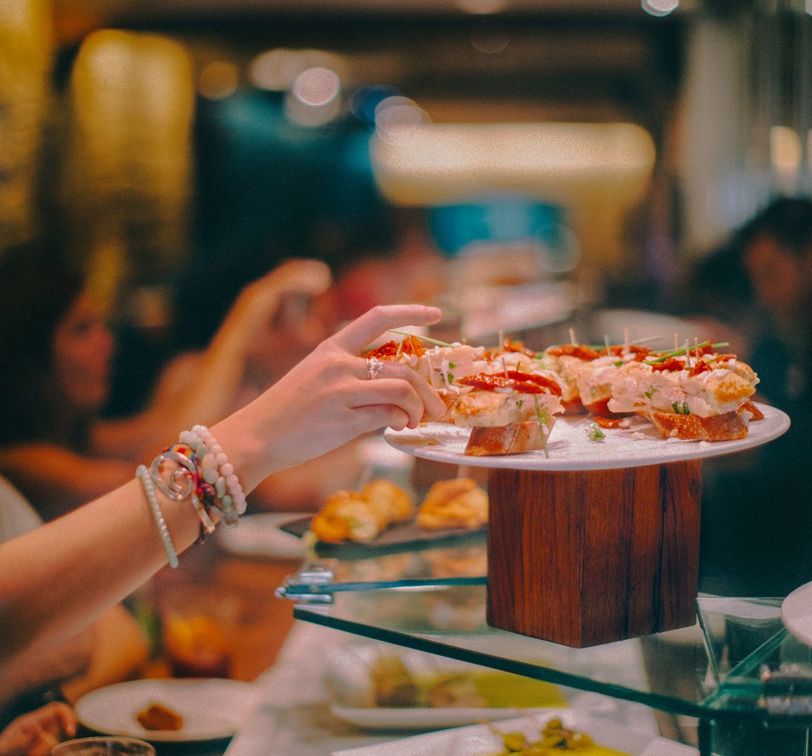 A person reaches for a piece of finger food on a decorative platter in a restaurant setting, with a blurred background of diners.