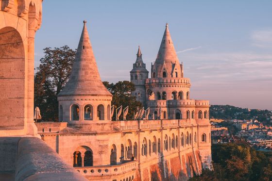 A castle-like structure with pointed towers, bathed in golden sunlight, set against a clear blue sky with lush greenery and a distant cityscape.