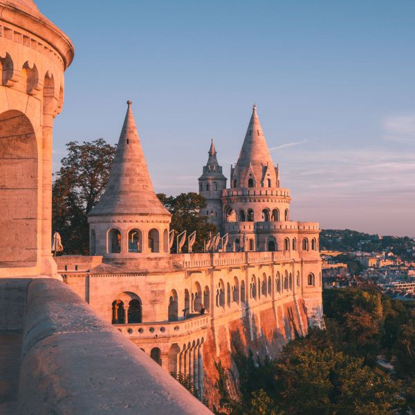 A castle-like structure with pointed towers, bathed in golden sunlight, set against a clear blue sky with lush greenery and a distant cityscape.