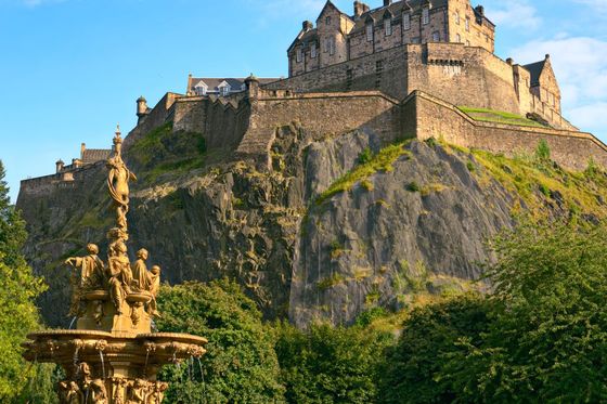 A historic castle sits atop a rocky hill surrounded by lush greenery, with an ornate fountain in the foreground and a clear blue sky above.