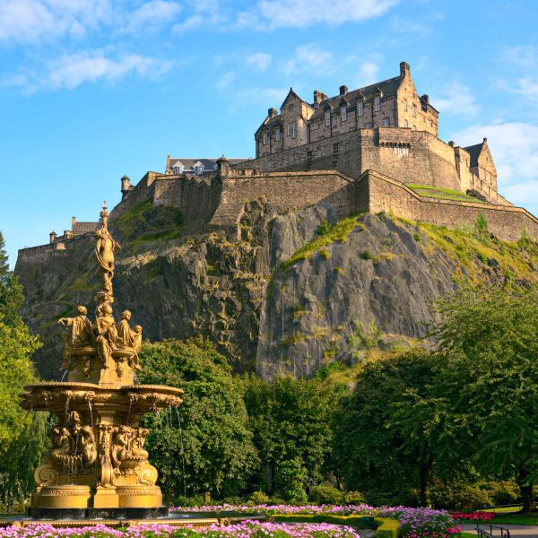 A historic castle sits atop a rocky hill surrounded by lush greenery, with an ornate fountain in the foreground and a clear blue sky above.