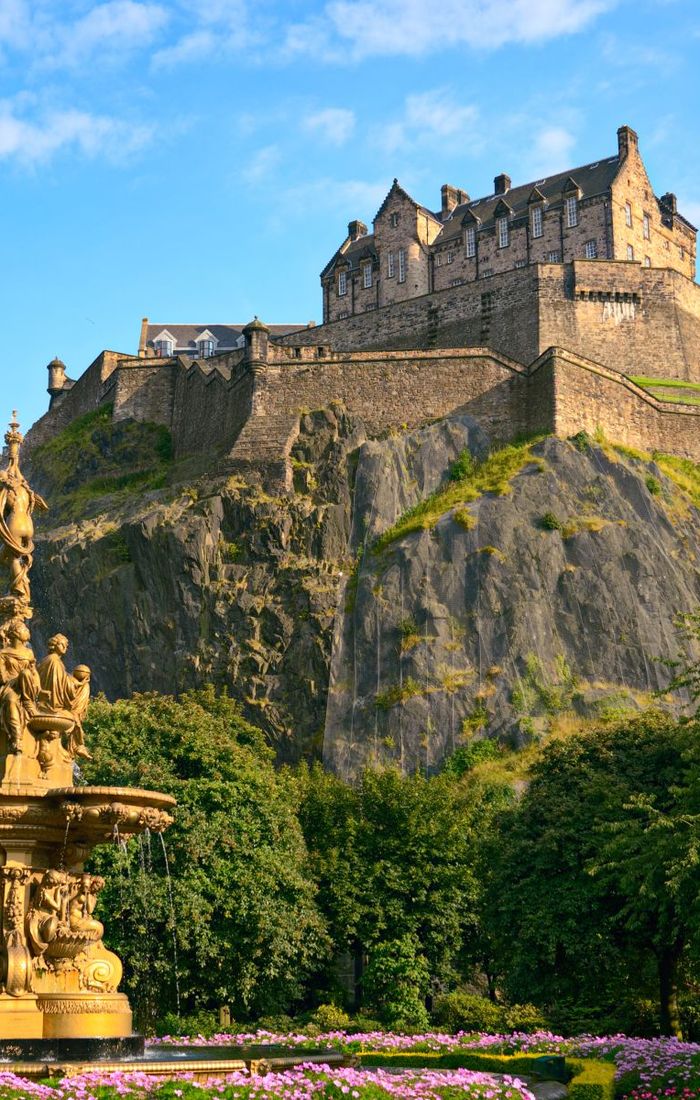 A historic castle sits atop a rocky hill surrounded by lush greenery, with an ornate fountain in the foreground and a clear blue sky above.