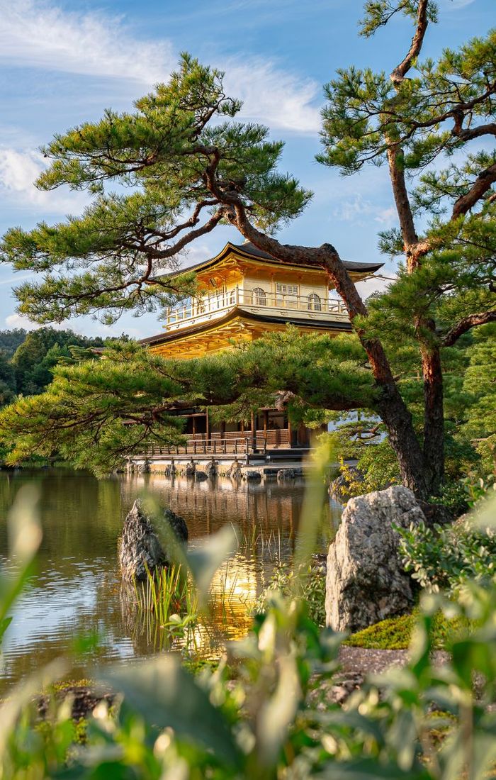 A golden pavilion surrounded by lush greenery and trees, reflected in a serene pond under a blue sky.