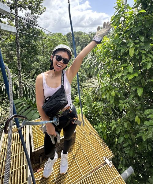 A woman in a helmet and harness, smiling and waving, stands on a yellow platform surrounded by lush green trees.