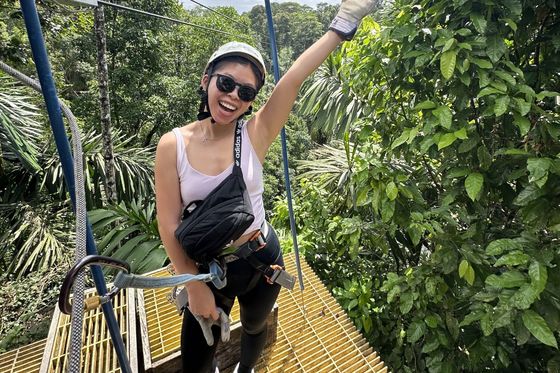 A woman in a helmet and harness, smiling and waving, stands on a yellow platform surrounded by lush green trees.
