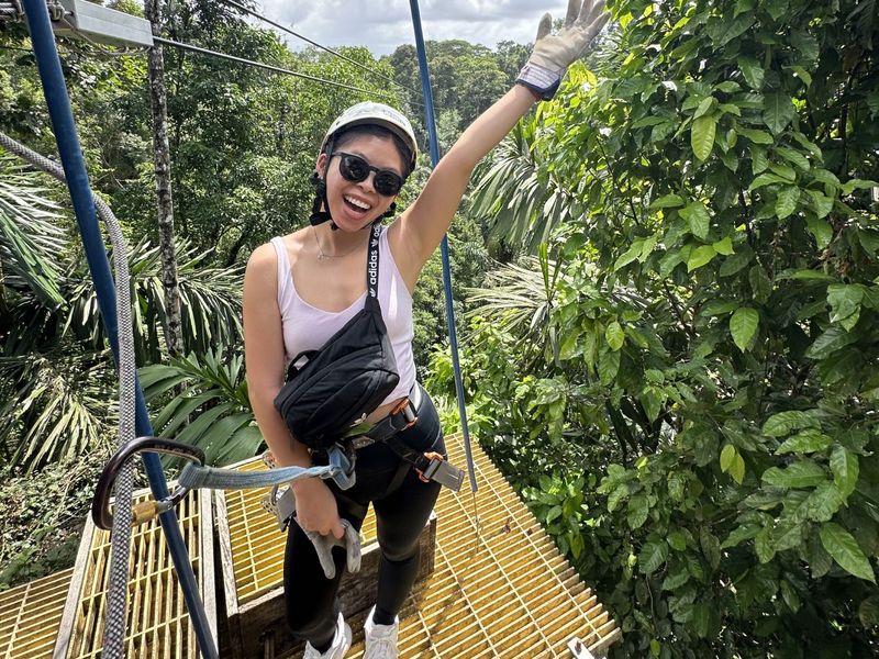A woman in a helmet and harness, smiling and waving, stands on a yellow platform surrounded by lush green trees.