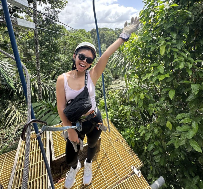A woman in a helmet and harness, smiling and waving, stands on a yellow platform surrounded by lush green trees.