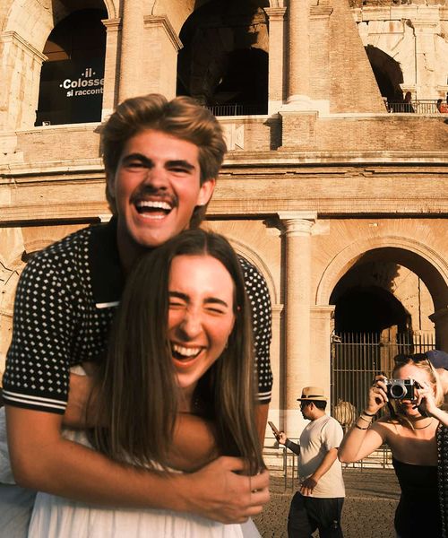 A couple laughs joyfully with the Colosseum in the background, as another person takes a photo of them.